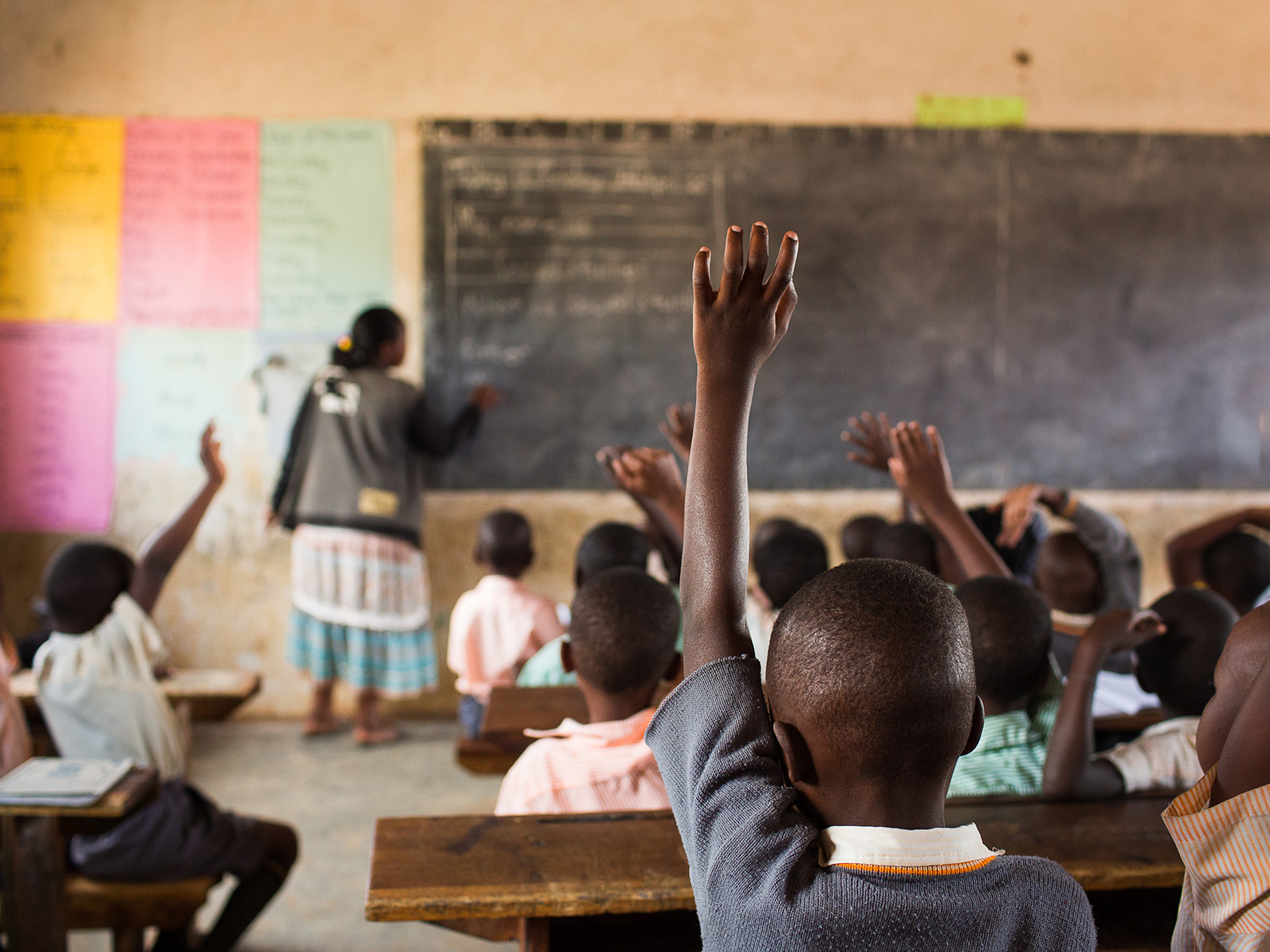 Children in a Ugandan classroom raise their hands to speak, while their teacher writes on the chalkboard